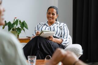 A smiling woman in a striped shirt takes notes in a notebook while seated comfortably in a counseling session with a couple, who are out of view.