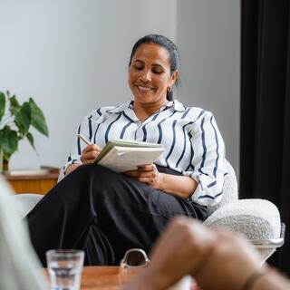 A smiling woman in a striped shirt takes notes in a notebook while seated comfortably in a counseling session with a couple, who are out of view.