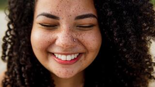 A joyful young woman with curly hair and freckles smiles brightly, exuding warmth and happiness. Her eyes are closed in delight.