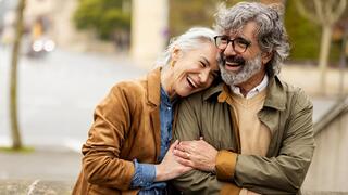 A joyful elderly couple stands together, sharing laughter and warmth, surrounded by a serene urban backdrop on a cloudy day.
