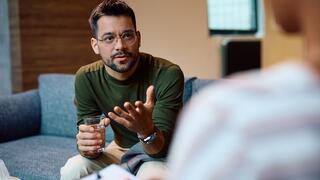 A man in a green sweater and glasses gestures while holding a glass of water, engaged in conversation.