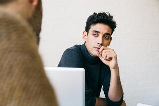 A young man, deep in thought, listens intently while resting his chin on his hand, engaged in a conversation with someone out of view.
