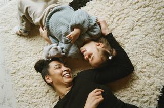 A woman and a young girl laugh while playing on a cozy rug, enjoying a playful moment.
