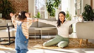A smiling woman sits with outstretched arms, joyfully welcoming a young child running towards her in a bright, cozy living room.