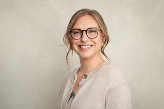 Holly from the Wellmind Team smiles warmly. She is wearing glasses and a beige cardigan against a soft, neutral backdrop. Her hair is styled in loose waves.