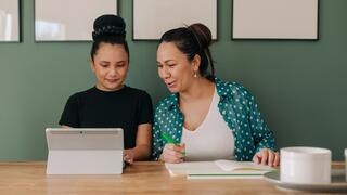 A woman and child engage in a learning session, with the child using a tablet while the adult assists her with a notebook.