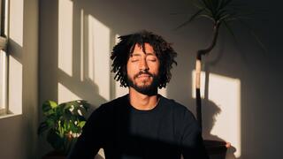 A man with curly hair sits peacefully with closed eyes, basking in soft sunlight, surrounded by plants and shadows.