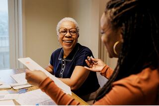 Two women share a joyful moment at a table, engaged in conversation with papers spread around them.