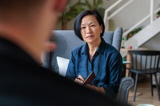 A woman in a blue shirt sits attentively in a modern, cozy setting, engaging in conversation with someone out of view.