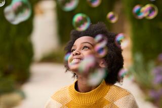 A smiling child with curly hair gazes upward in delight, surrounded by floating bubbles in a vibrant, green outdoor setting.
