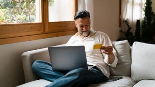 A man smiles while seated on a couch, holding a card in one hand and using a laptop on his lap, surrounded by natural light.