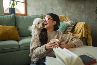 A young woman enjoys a cozy moment on a couch, holding a mug, while a small dog playfully rests on her shoulder, creating a warm atmosphere.