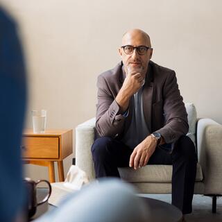 A man in glasses sits thoughtfully on a couch, dressed in a blazer, engaged in a conversation.