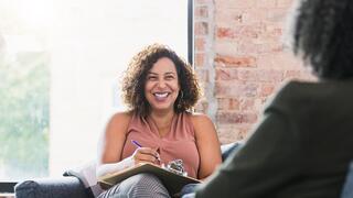 A smiling woman takes notes while engaged in a conversation, sitting comfortably in a cozy, well-lit space with a brick wall background.