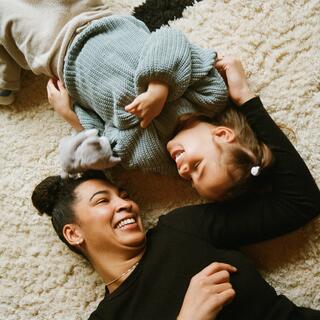 A woman and a young girl laugh while playing on a cozy rug, enjoying a playful moment.