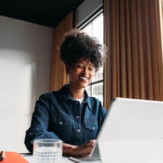 A woman with curly hair smiles while working on a laptop, enjoying the bright ambiance of a cozy, modern workspace.