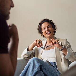 A smiling woman in a light jacket sits comfortably, holding her glasses, engaging in conversation with a man nearby.