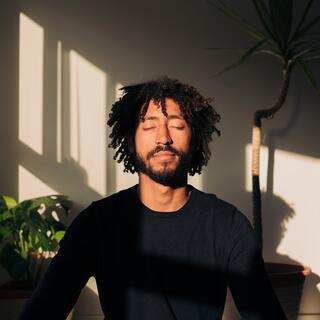 A man with curly hair sits peacefully with closed eyes, basking in soft sunlight, surrounded by plants and shadows.