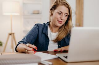 A woman with long hair sits at a desk, engaging thoughtfully with content on her laptop while taking notes.